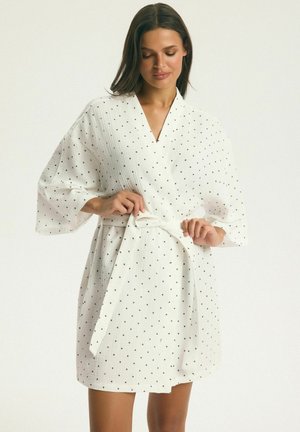 Žena si upravuje opasok bieleho, krátkeho župana s bodkami a širokými rukávmi na jednoduchom pozadí.