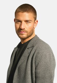 Grå stickad blazer över en svart skjorta, med en kavajkrage och lös passform. Kort hår och lätt skäggstubb fulländar looken.