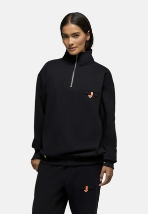 Femme portant un pull noir à fermeture éclair quart et un pantalon assorti avec de petits détails orange et boutons sur les poches, debout devant un fond uni.