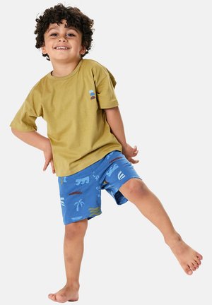 Smiling child with curly hair wearing mustard t-shirt and blue shorts with tropical patterns lifts one leg while standing barefoot.