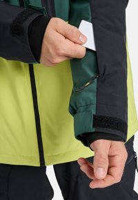 Jacket featuring a bright yellow body and dark green sleeves, with a textured black Velcro strap on the cuff and a zippered pocket for cards.