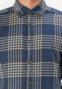 Karierte Button-up-Hemd in Marineblau und Creme mit blauen Akzenten. Hergestellt aus einem strukturierten Stoff mit einem feststehenden Kragen und braunen Knöpfen.
