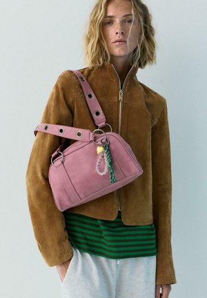 Blondína v hnedej semišovej bunde a zelenej pruhovanej košeli, nesie ružovú kabelku s dekoratívnymi príveskami cez rameno.