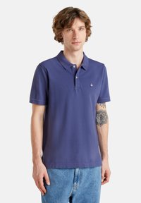 Camisa polo de color azul marino con un placket de tres botones y un pequeño logo en el pecho, hecha de algodón con una textura suave, combinada con unos jeans de mezclilla claros.