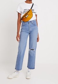 Persona vistiendo una camiseta gráfica blanca, jeans rotos de color azul claro, zapatillas blancas y un bolso bandolera amarillo mostaza contra un fondo liso.