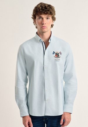 Camisa de botones a rayas azul claro con mangas largas, que presenta un escudo bordado en el lado izquierdo del pecho y un detalle en el interior del cuello de contraste.