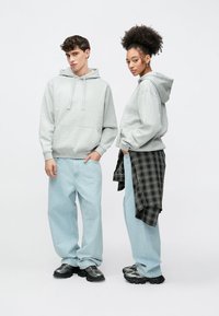 Grå hettegensere med frontlommer, kombinert med lyseblå vide jeans. En person har en svart rutete skjorte knyttet rundt midjen.