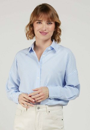 Femme aux cheveux courts et ondulés portant une chemise à boutons rayée bleu clair et blanc et un pantalon blanc, souriante avec les mains doucement jointes.