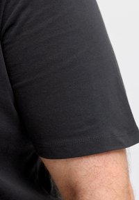 Κοντινό πλάνο του βραχίονα ενός ατόμου που φοράει κοντομάνικη μαύρη βαμβακερ ή μπλούζα, πάνω σε απλό ανοιχτόχρωμο φόντο.