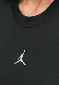 Κοντινή λήψη ενός μαύρου πουκαμίσου με ένα μικρό λευκό σιλουέτα λογότυπο ενός ατόμου που πηδάει και κρατάει μια μπάλα μπάσκετ κοντά στην περιοχή του στήθους.