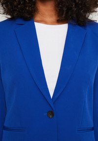 Blå kavaj med en slät textur, snedställda slag och en enda framknapp. Bärs över en vit, ribbad topp. Sidofickdetalj synlig.