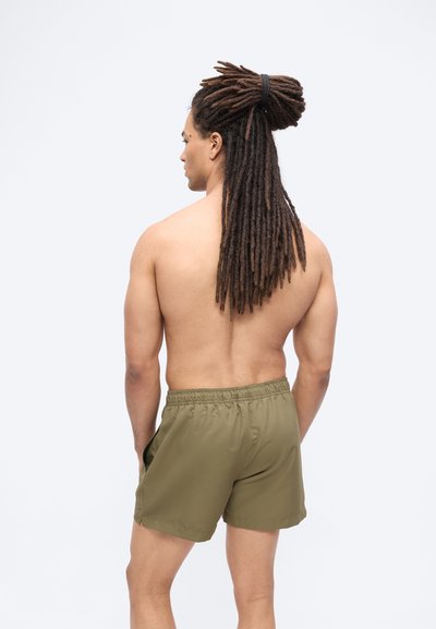 Άνδρας με μακριά dreadlocks δεμένα, χωρίς μπλούζα, φορώντας χακί σορτς, στέκεται με τα χέρια στις τσέπες, γυρισμένος με την πλάτη σε απλό φόντο.