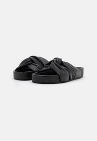 Černé skluzavky s uzlovým designem popruhu, vyrobené z hladkého, flexibilního materiálu s plochou podrážkou a minimalistickým vzhledem.
