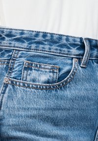 Μπλε ζώνη από τζιν και μπροστινή τσέπη με θηλιά για ζώνη, φορεμένα πάνω από λευκό ριμπ ύφασμα.