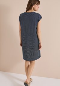Femme de dos portant une robe bleu marine mi-longue à manches courtes avec de fines rayures horizontales et des sandales beige à enfiler.