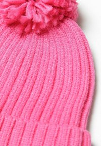Ráncos kötött rózsaszín sapka, tetején bolyhos pom-pommal. Az anyag puhanak tűnik, textúrája bolyhos, ami melegséget és kényelmet ad.