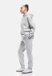 Jasnoszary hoodie z przednią kieszenią, kapturem na sznurki i szczegółami tekstowymi. Połączony z dopasowanymi dresami oraz białymi trampkami z brązowymi podeszwami.