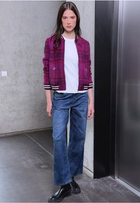 Veste bomber à carreaux bordeaux avec des poignets rayés blancs et noirs, associée à un jean large bleu et des bottes noires chunky. T-shirt blanc simple en dessous.