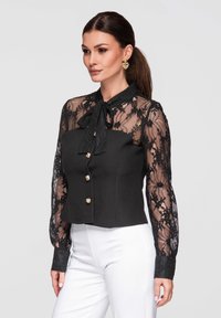 Femme portant un chemisier en dentelle noire à manches longues avec col à nœud et boutons dorés, assorti à un pantalon blanc taille haute, tournée légèrement vers la droite.