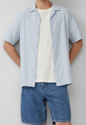 Uomo che indossa una camicia azzurro chiaro a maniche corte abbottonata sopra una t-shirt bianca e pantaloncini di jeans blu, in piedi con le mani dietro la schiena.