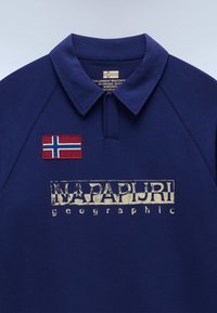 Tmavě modré polo tričko s límečkem, s červeně, bíle a modře zbarvenou obdélníkovou vlajkovou nášivkou a opraným bílým nápisem "NAPAPIJRI geographic" na přední straně.