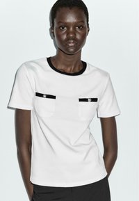 White short-sleeve t-shirt with black trim and two chest pockets. Each pocket has a silver button detail, made from smooth fabric.