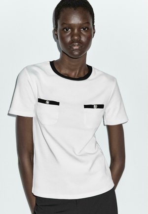 White short-sleeve t-shirt with black trim and two chest pockets. Each pocket has a silver button detail, made from smooth fabric.