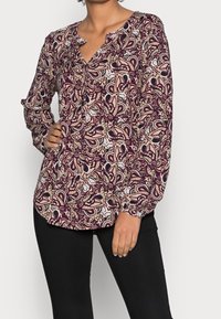 Blusa a maniche lunghe con un motivo paisley in viola e beige, con scollatura a V e dettagli con bottoni. Tessuto morbido, vestibilità leggermente ampia.