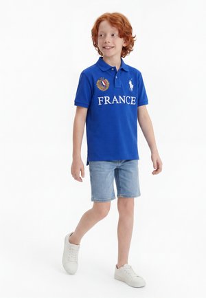 Αγόρι με κόκκινα μαλλιά που φοράει μπλε πόλο μπλούζα με την επιγραφή «FRANCE» και έμβλημα, τζιν σορτς και λευκά αθλητικά παπούτσια, περπατώντας και χαμογελώντας σε λευκό φόντο.