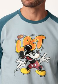 Camiseta de color azul claro con mangas raglán en color teal, que presenta un personaje de dibujos animados, Mickey Mouse, y un texto en negrita que dice "Estoy Perdido" en naranja y amarillo.