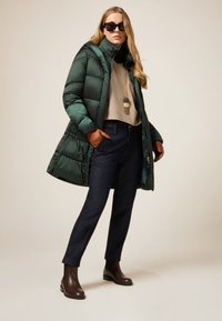 Abrigo acolchado verde, suéter beige, jeans oscuros, guantes marrones y botines. Los accesorios incluyen un collar largo con colgantes circulares.