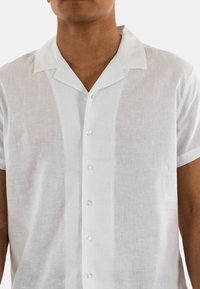Camisa de lino blanca de manga corta con cuello tipo camp, cierre de botones y botones transparentes. La tela tiene un aspecto ligeramente texturizado.