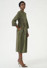 Olijfgroene, knoopsluiting jurk met driekwartmouwen, geplooide taille en zijzakken. Draagt met beige enkelsandalen met enkelband.