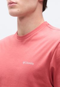 Κοντινό πλάνο ενός ατόμου που φοράει ροζ μπλούζα Columbia με το λογότυπο ορατό στο στήθος, πάνω σε ένα απλό φωτεινό φόντο.