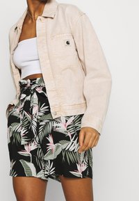 Světle béžová denimová bunda s přední kapsou, přes bílý pruhovaný crop top. Černé šortky s tropickým vzorem listů a mašlí v pase.