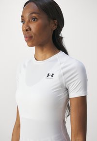 Hvid træningsshirt med korte ærmer, der har et tætsiddende design og glat tekstur. Har et sort Under Armour-logo på venstre side af brystet.