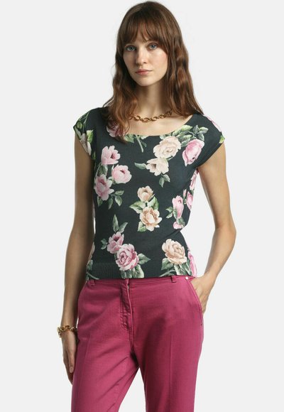 Femme aux cheveux bruns ondulés portant un haut noir à manches courtes avec motif floral, un pantalon rose, un collier chaîne en or et un bracelet assorti, posant avec une main dans la poche.