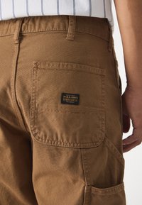 Bruna cargo-byxor med en bakficka och en svart etikett. Tyget är hållbart med en slät yta och synliga sömnadsdetaljer.