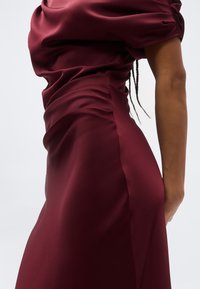 Kvinne iført en tettsittende, kortermet vinrød satengkjole, vist fra siden mot en ensfarget lys bakgrunn.