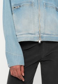 Ανοιχτού γαλάζιου τζιν μπουφάν με φερμουάρ, δύο μπροστινές τσέπες και ελαφρώς κοντή γραμμή. Φορεμένο πάνω από μαύρο ριγέ παντελόνι.