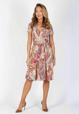 Frau steht, trägt ein knielanges, buntes Kleid mit Paisley-Muster, kurzen Ärmeln und braunen High Heels.