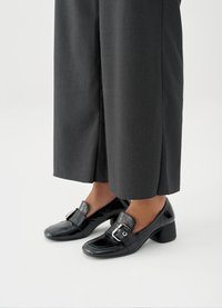 Μαύρα παπούτσια από δερματίνη με τετράγωνη μυτερή μύτη, χαμηλό χοντρό τακούνι και διακοσμητική αγκράφα στο πλάι, συνδυασμένα με γκρι παντελόνια φαρδιάς γραμμής.