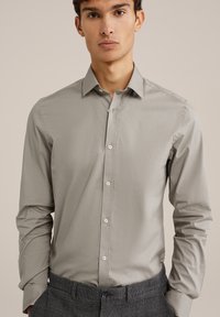 Chemise à manches longues gris clair avec un col pointu, fermeture à boutons et tissu texturé. Associée à un pantalon à carreaux gris.