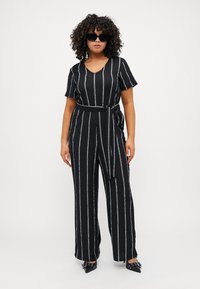 Black jumpsuit with vertical white and colored stripes, short sleeves, V-neck, and a tied waist belt. Worn with oversized black sunglasses.