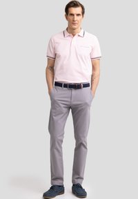 Camisa polo rosa claro con cuello a rayas en negro y blanco, combinada con pantalones gris y zapatos azules. Muestra un corte relajado y un estilo casual.