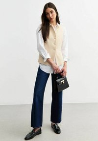Chaleco de corte entallado en color beige con botones, camisa blanca debajo, jeans de pierna ancha en azul oscuro, bolso texturizado negro y mocasines negros.