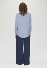Persoon met krullend haar, gekleed in een lichtblauw langemouwen bovenstuk en een brede navy broek, staat met de rug naar de camera tegen een effen achtergrond.
