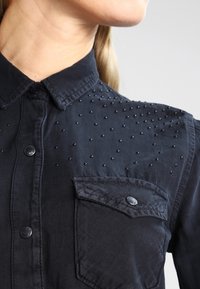Femme portant une chemise noire à boutons avec de petites perles noires sur l'épaule et une poche poitrine boutonnée, faisant face à droite.