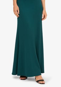 Tyrkysové dlouhé šaty z lehké tkaniny s volným střihem. Nosené s černými skluznými sandály, které vystavují otevřenou špičku a minimalistický styl.