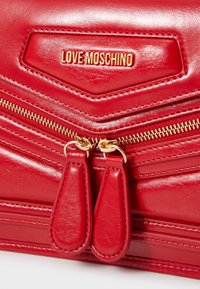 Röd konstläderhandväska med chevronmönster, guldzipper med dubbla dragkedjor och en guldlogotavla präglad på framsidan.